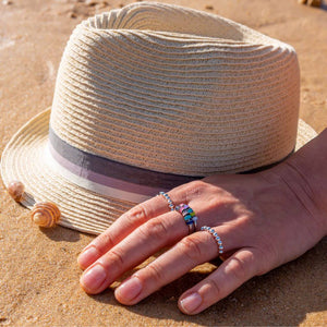 Woman's hand on sand wearing 3 of the best anxiety rings in Australia next to a straw hat