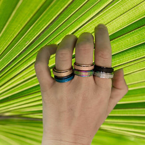Woman's hand wearing six spinner rings silver, gold, rose gold, blue, black on green background